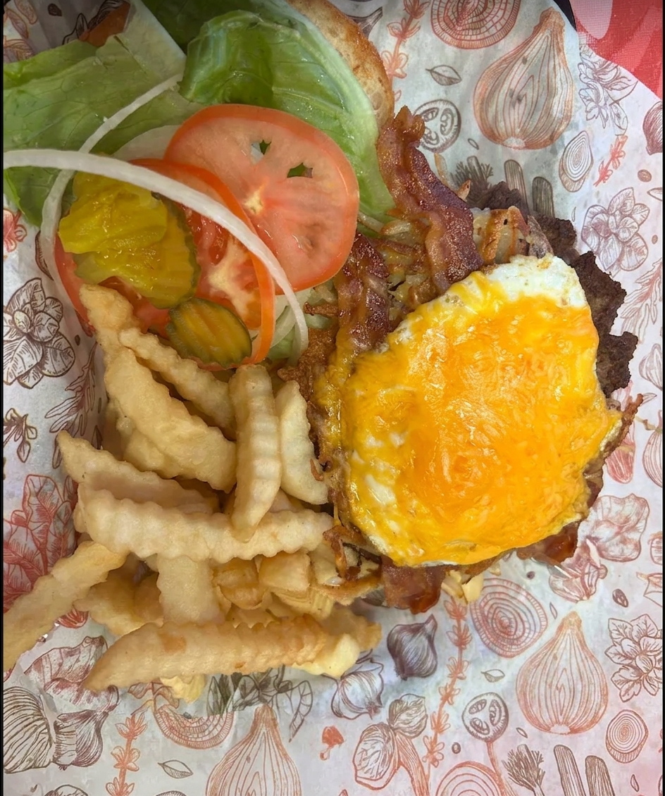 A burger topped with a fried egg over crinkle fries in a paper-lined basket